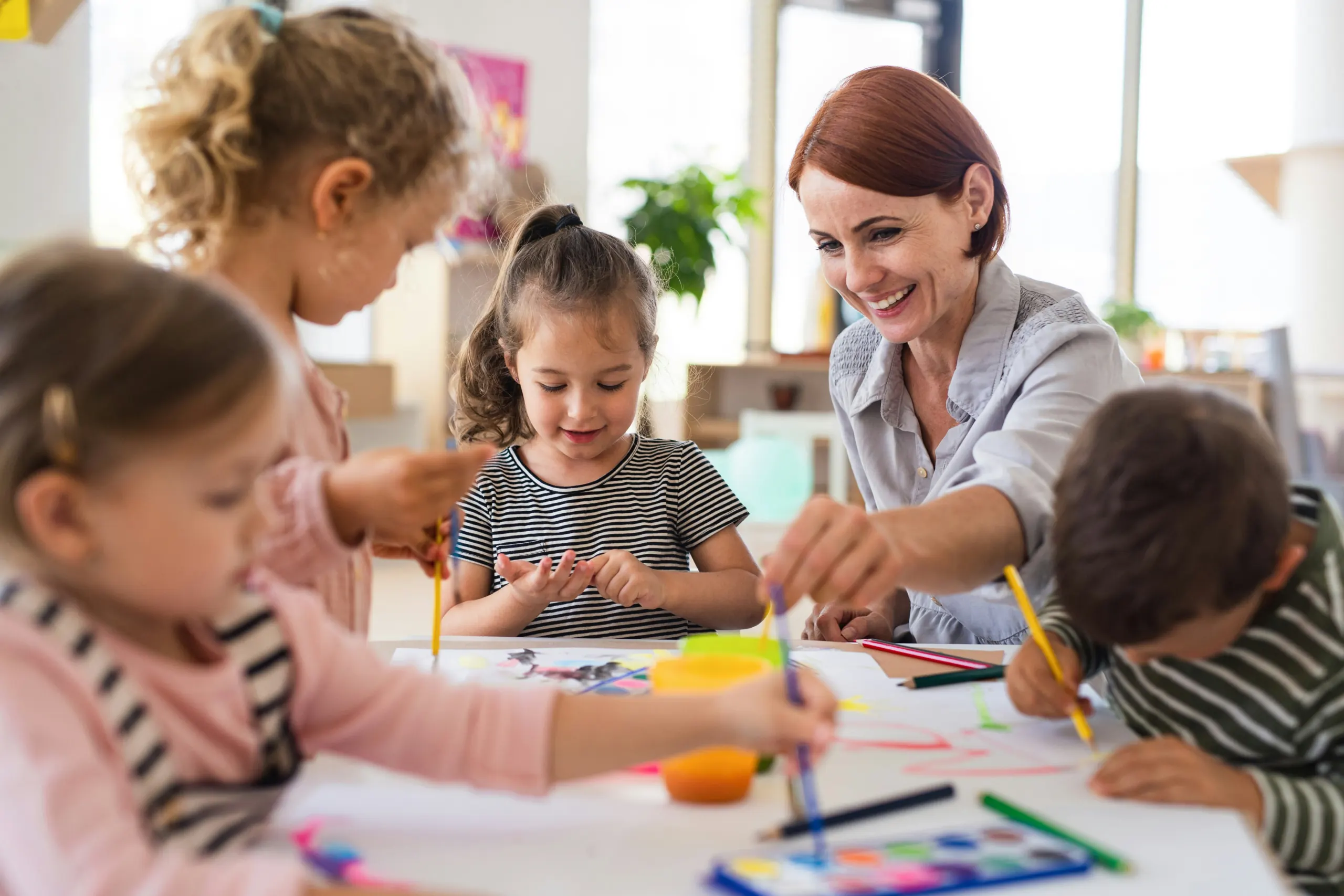 A teacher leaning over a table of young children painting and drawing together in a bright classroom